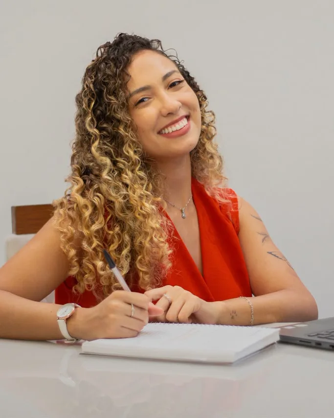 Psicóloga Isabella Lacerda sorrindo, sentada à mesa com um caderno e o notebook